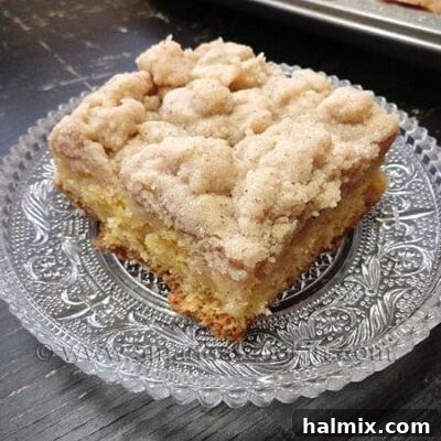 A close up photo of a square of crumb cake on a clear plate, showcasing its golden-brown streusel topping.