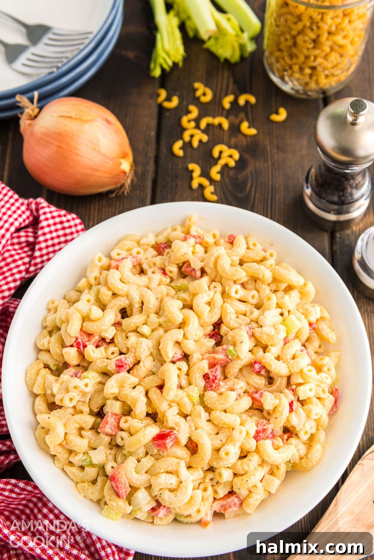 Creamy Macaroni Salad with Fresh Vegetables A rustic table setting featuring a large bowl of macaroni salad, with fresh bell peppers, onion, dried elbow macaroni, and stacked plates in the background.