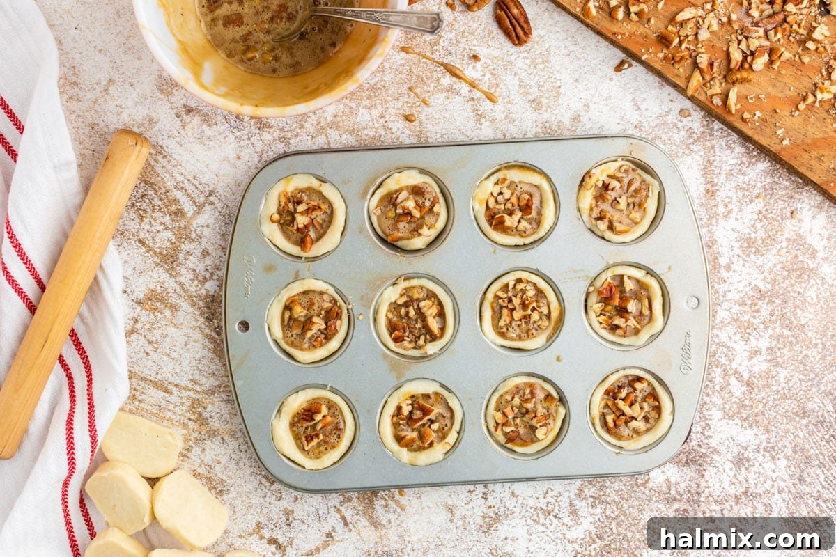 Unbaked pecan tassies in a mini muffin tin, topped with chopped pecans, ready to go into the oven.