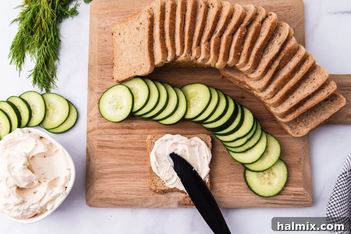 Refreshing Cucumber Finger Sandwiches 5 Close-up showing a hand spreading cream cheese on a slice of cocktail rye bread, demonstrating the first step in making mini cucumber sandwiches.