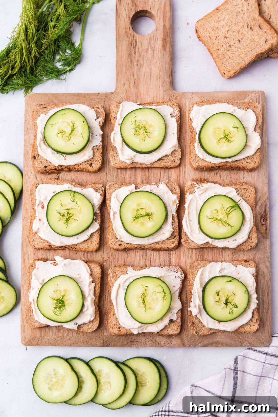 Refreshing Cucumber Finger Sandwiches 7 A wooden board filled with open-faced mini cucumber sandwiches, garnished with dill and ready for serving.
