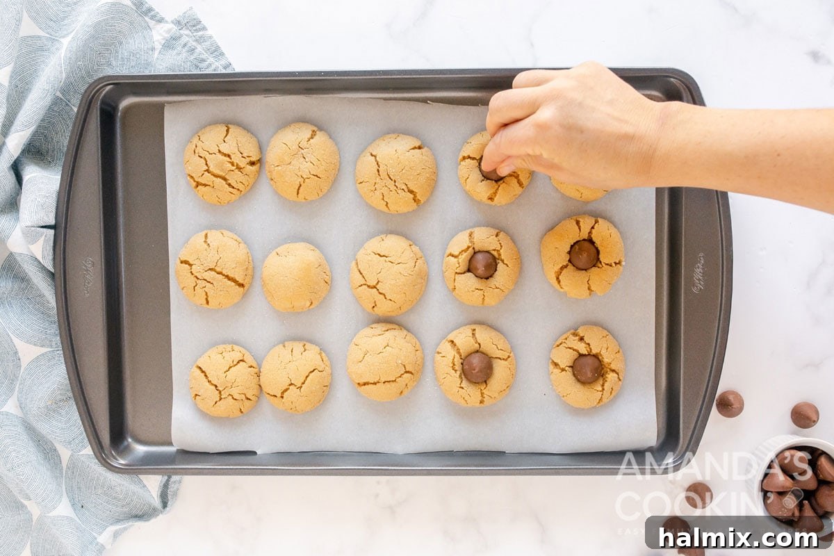 Gently pressing a Hershey's Kiss into the warm center of a freshly baked peanut butter cookie on a baking sheet.