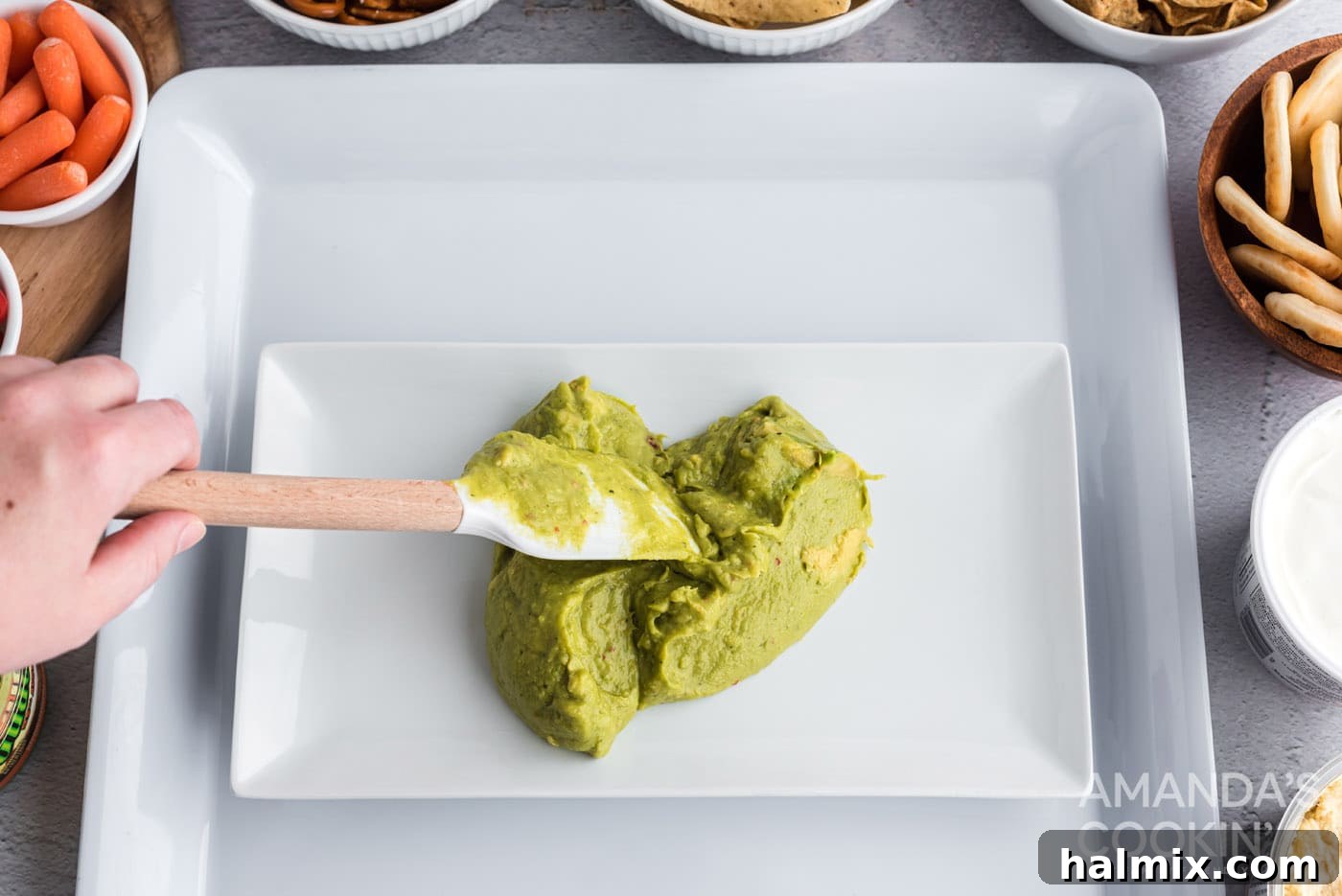 Hands carefully spreading vibrant green guacamole onto a smaller platter, forming the base of the football field design.