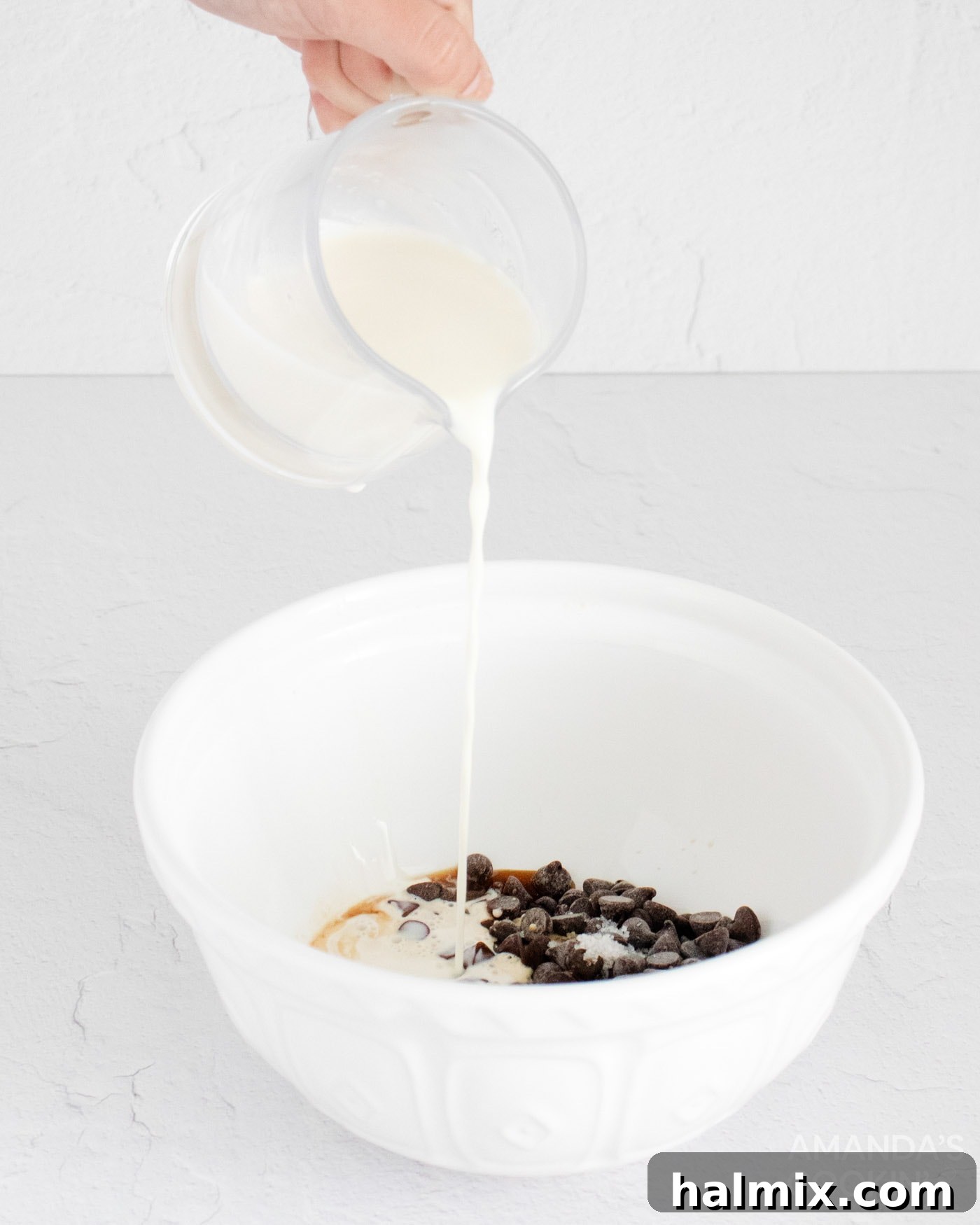 Warm heavy cream being poured into a large bowl filled with semi-sweet chocolate, espresso, butter, vanilla, and sea salt, ready to be whisked.