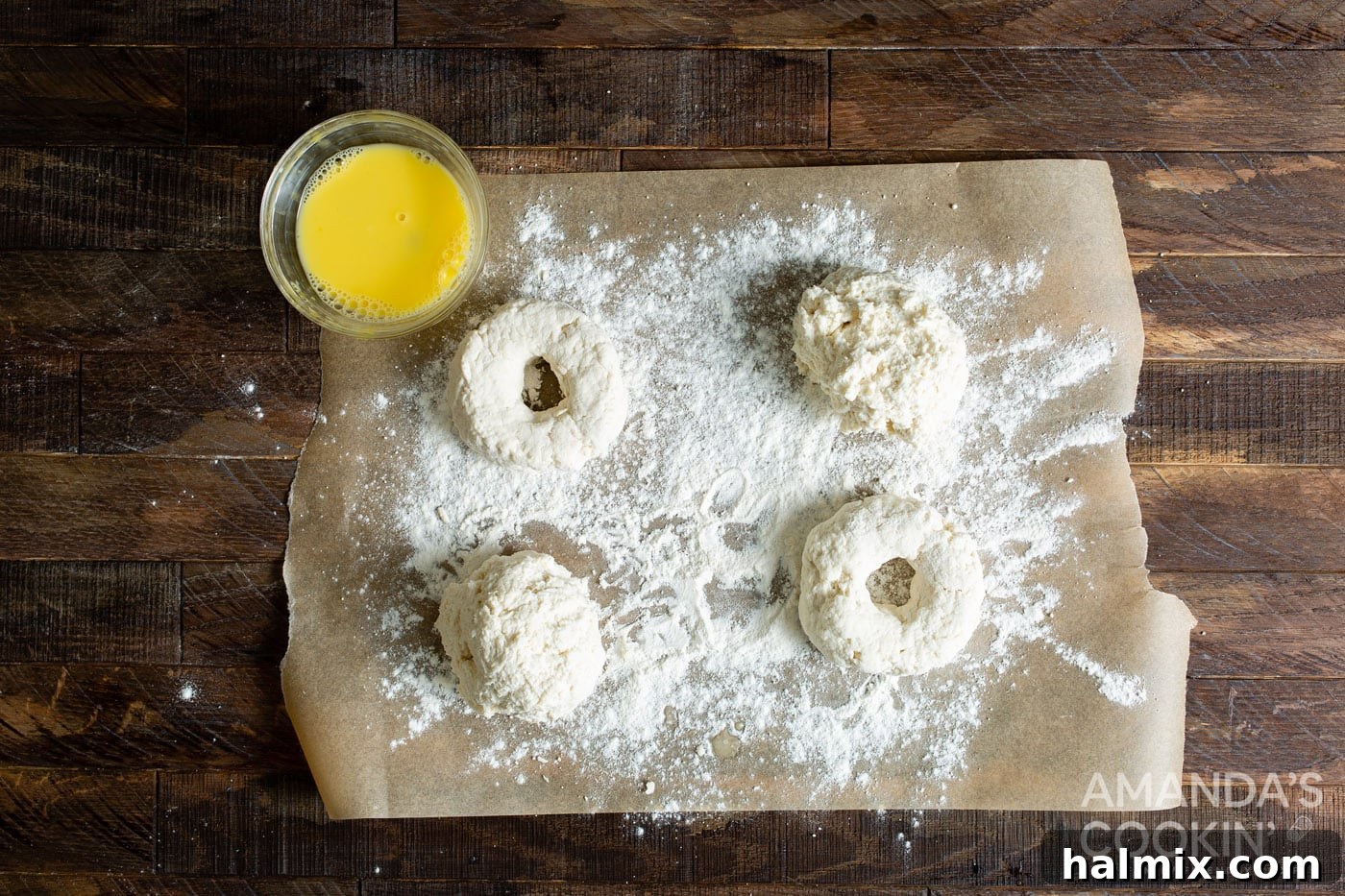 Raw bagel dough patties on parchment paper, with a thumb pressing through to create the center hole.