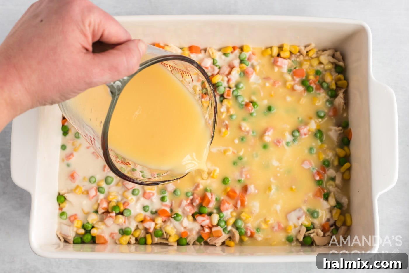 The whisked broth and soup mixture being slowly poured over the flour mixture layer in the casserole.