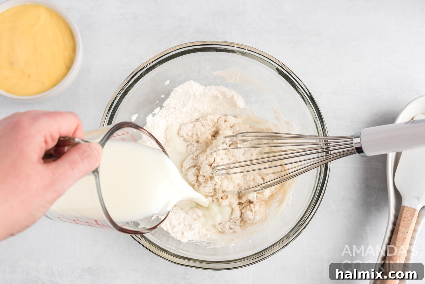 A whisk blending milk into dry ingredients (flour, baking powder, salt) in a mixing bowl.