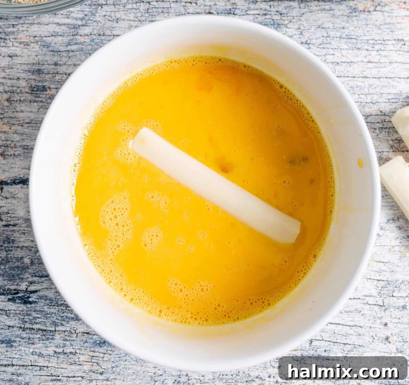 A hand dipping a string cheese stick into a bowl of whisked egg wash.