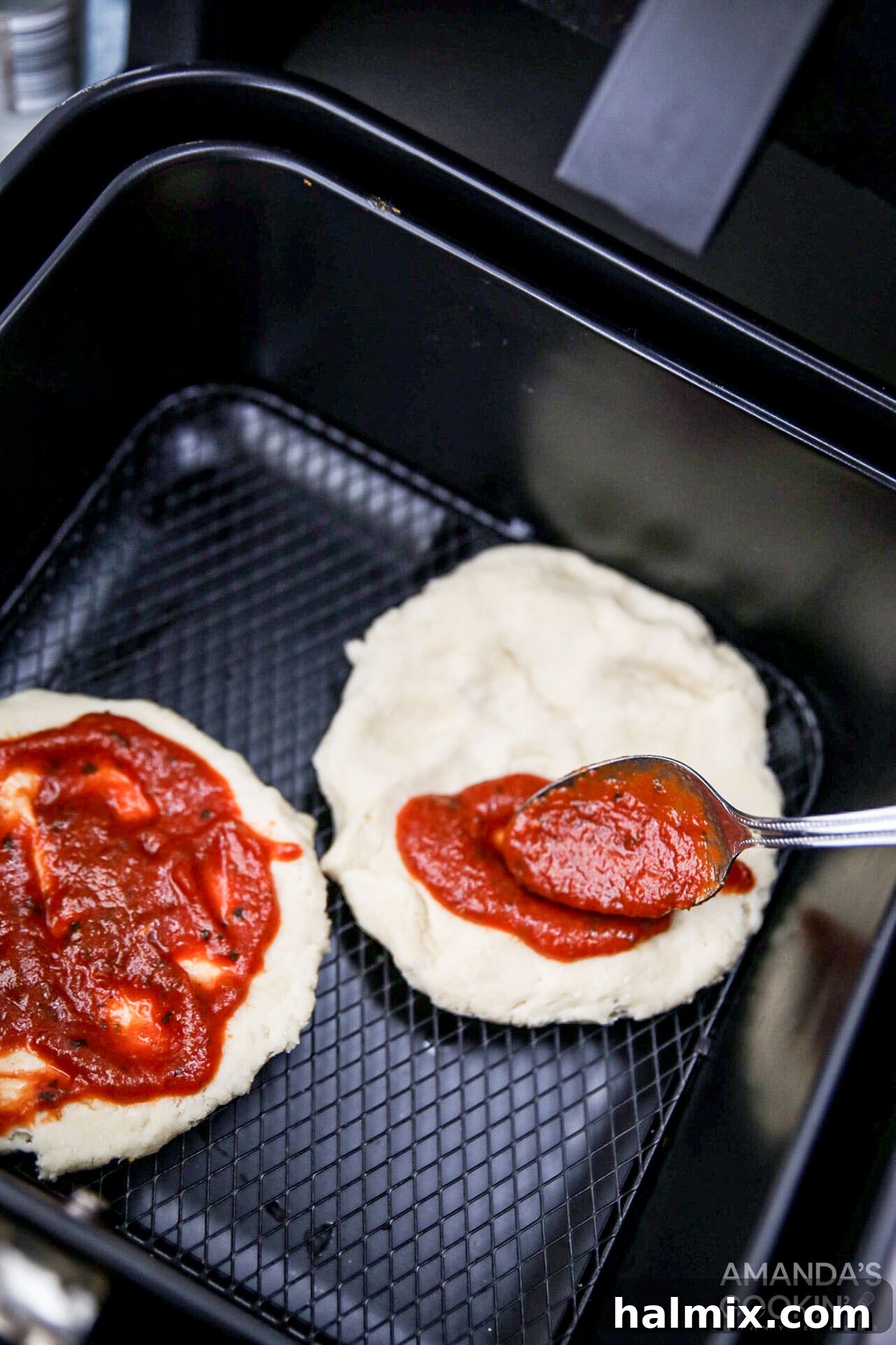 A hand spreading a generous layer of red pizza sauce onto a raw pizza crust already positioned in the air fryer basket.