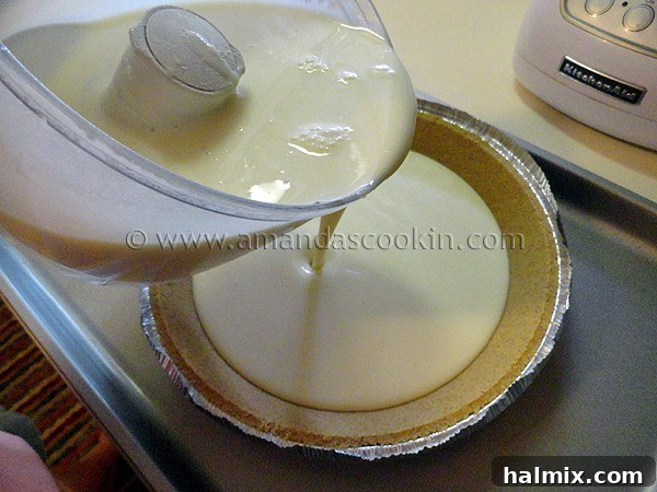 A photo of the ingredients for lemonade cheesecake in a food processor being poured into a graham cracker pie crust.