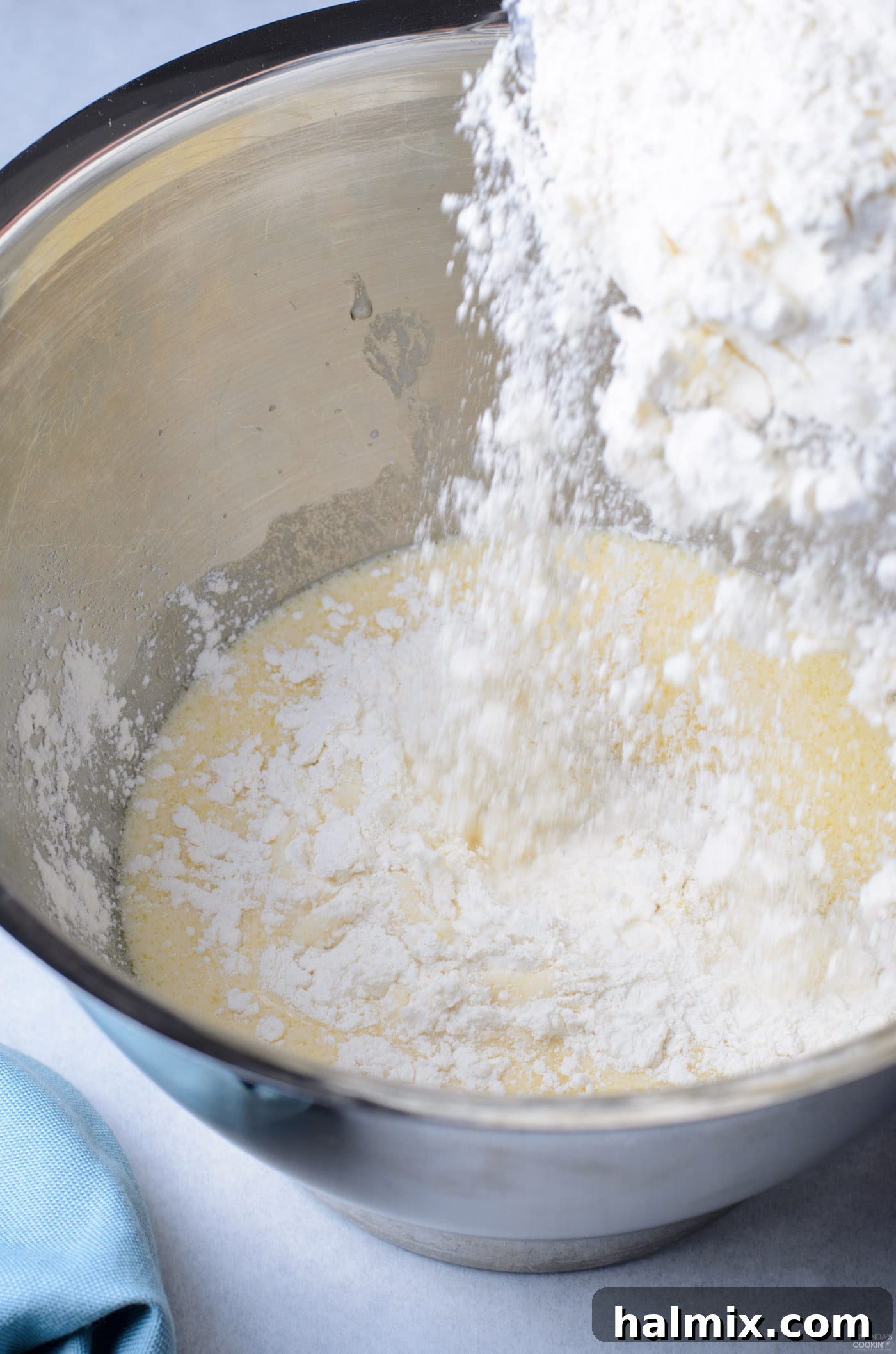 A stream of white flour being poured into the yeast mixture in a large mixing bowl.