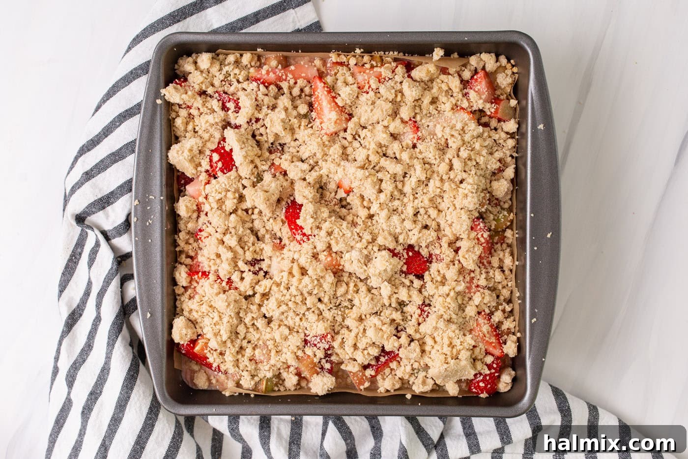 The final layer of oat crumble topping evenly spread over the strawberry rhubarb filling in the pan, ready for its journey to golden perfection in the oven