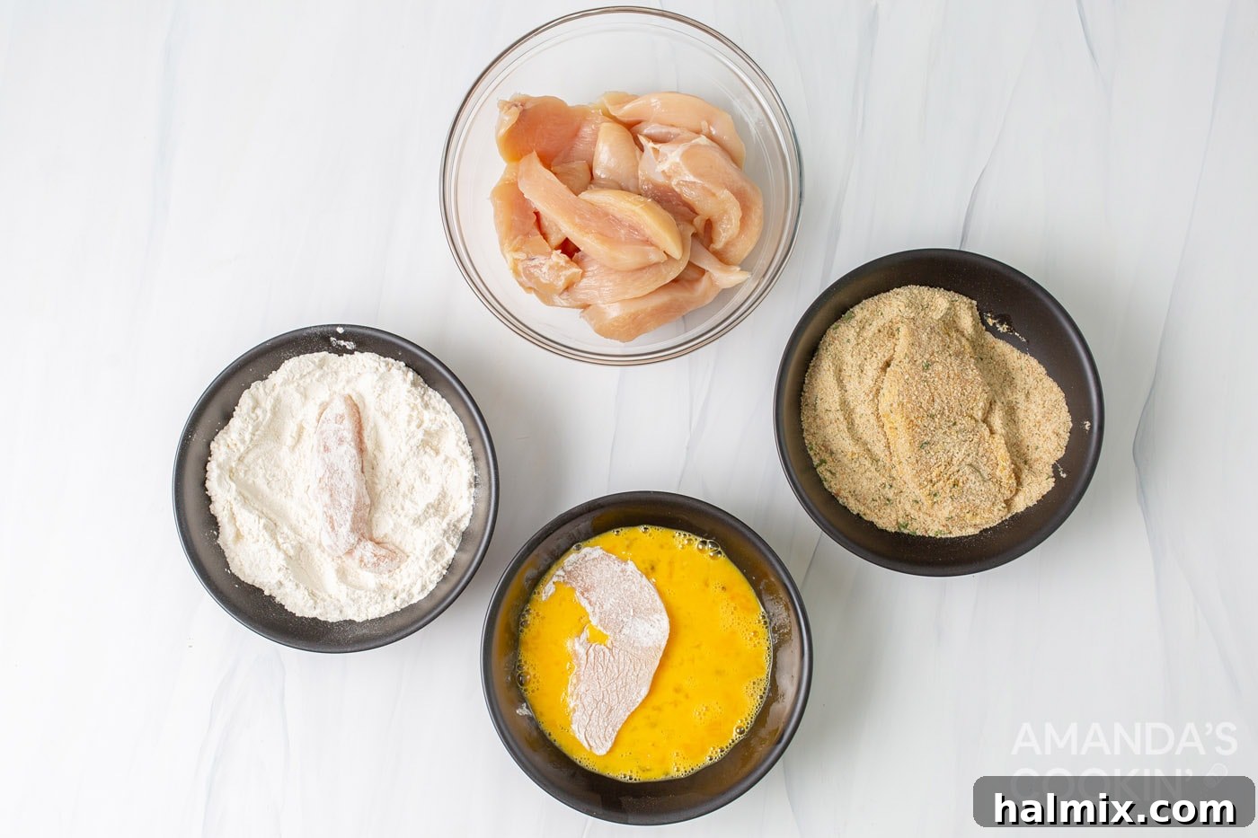 Bowls arranged for dredging: flour, eggs, seasoned breadcrumbs, and raw chicken tenders ready for coating