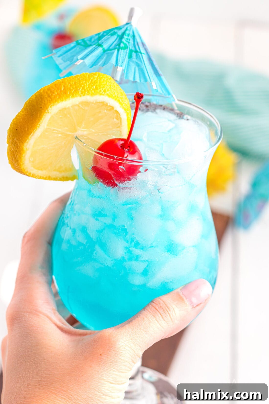 Azure Escape 3 Holding a freshly prepared Blue Lagoon Cocktail with a tropical background.