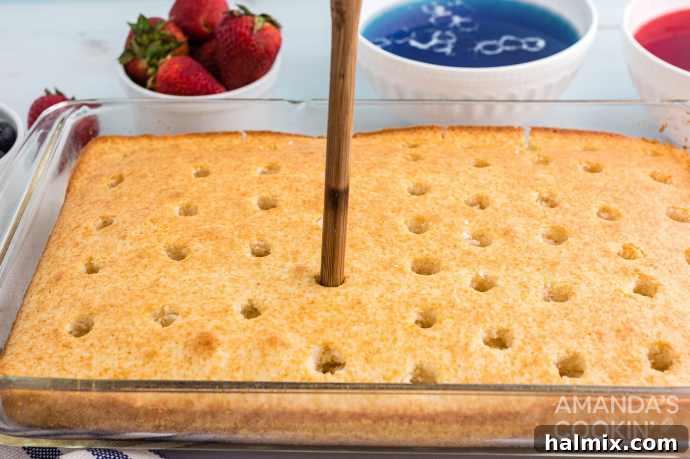 A close-up of a white cake being poked with the handle of a wooden spoon to create holes for the Jell-O.