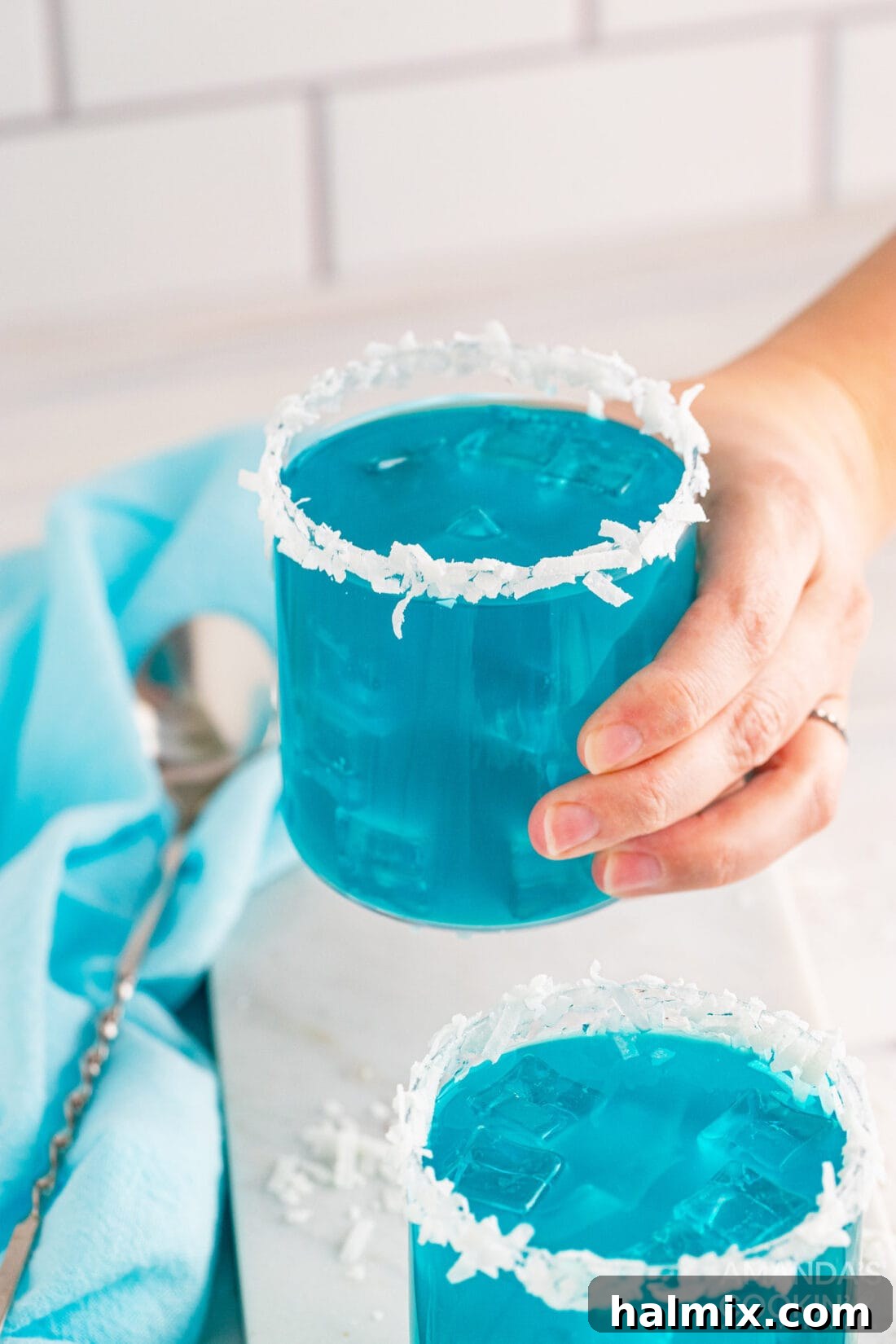 Neptune's Kiss Cocktail 3 A hand holding a frosty glass of Ocean Water Cocktail, garnished with a slice of orange and a tiny umbrella, against a blurred beach background.