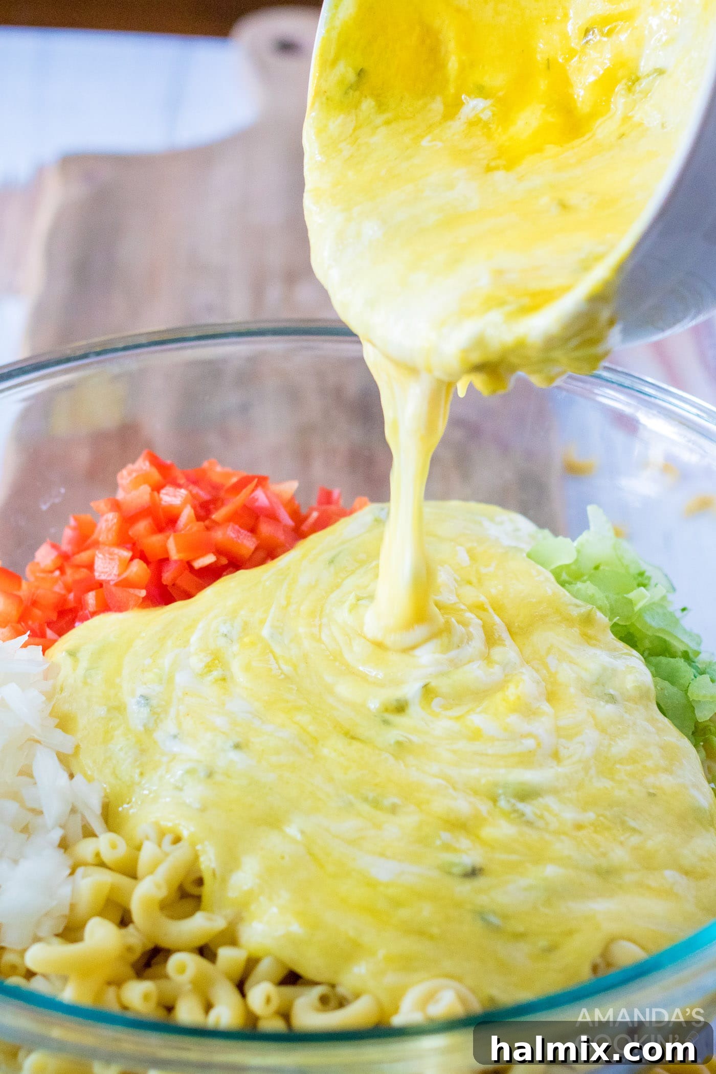 A large mixing bowl containing cooked elbow macaroni, chopped vegetables, and hard-boiled eggs, with the creamy dressing being poured over them, ready to be mixed for a vibrant Amish Macaroni Salad.