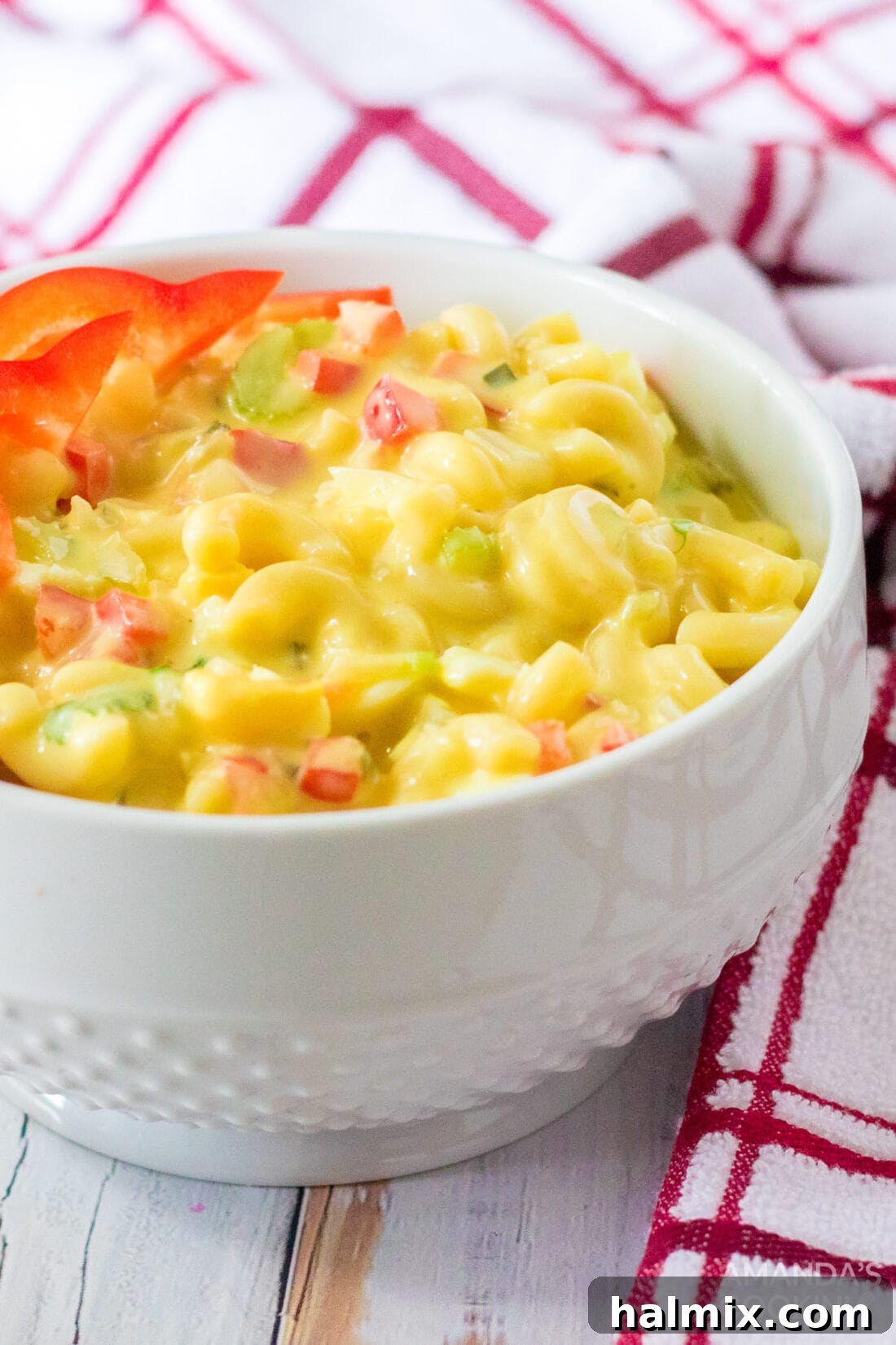 A small, elegant white bowl brimming with homemade Amish Macaroni Salad, featuring tender macaroni, creamy dressing, and colorful vegetables, presented against a soft, blurred background, highlighting its delectable appeal.