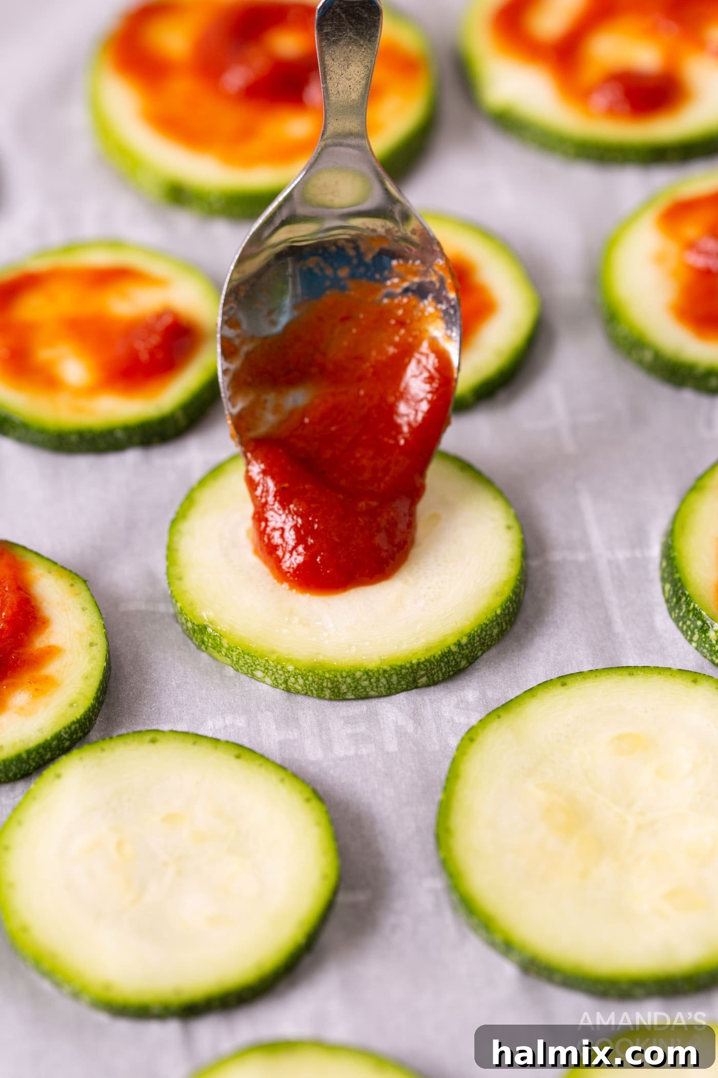 Spoon applying pizza sauce onto a zucchini slice