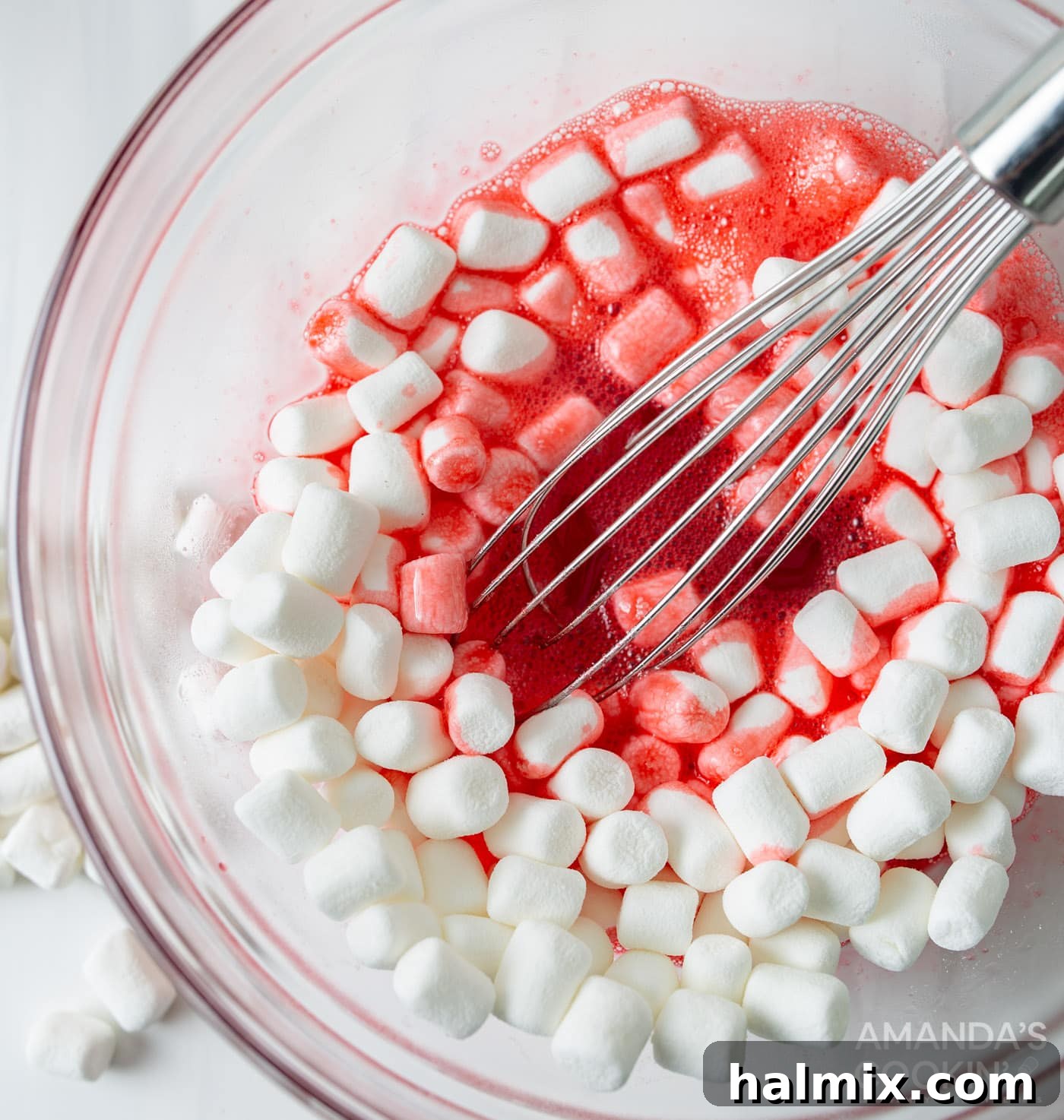 A whisk stirring mini marshmallows into dissolved red jello in a clear glass bowl, showing the mixture turning smooth and vibrant.