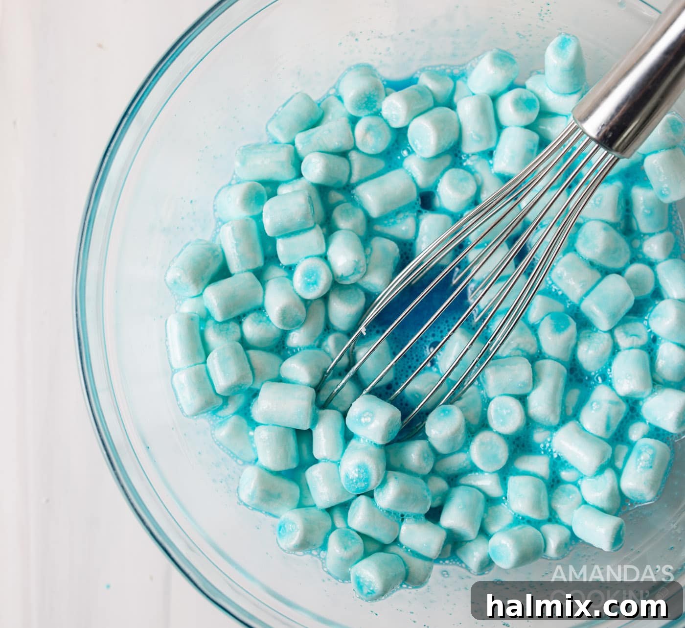 A blue jello and marshmallow mixture being whisked in a glass bowl, next to a box of berry blue Jell-O mix, highlighting the preparation of the second color.