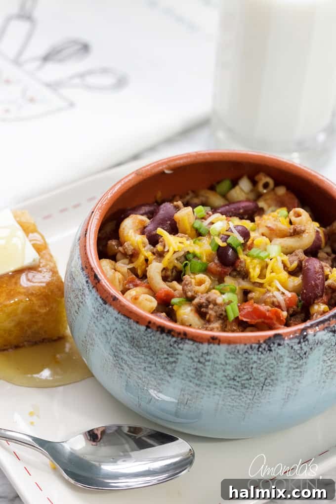 Chili Mac is a delicious combination of ground beef, beans, tomatoes, chili seasoning, and macaroni. This chili mac recipe makes a hearty dinner or a delicious cup of warmth for friends watching football! #chilimac #chili #groundbeef #beans #macaroni #footballrecipes #comfortfood #winterfood #chili A steaming bowl of homemade chili mac, garnished with fresh green onions and shredded cheese, ready to be enjoyed.