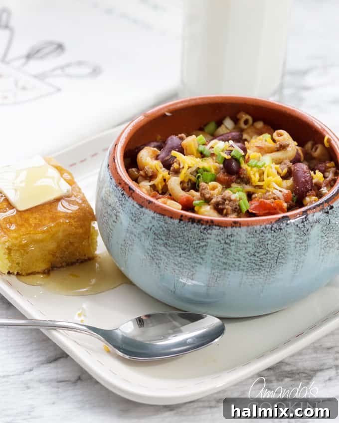 Chili Mac is a delicious combination of ground beef, beans, tomatoes, chili seasoning, and macaroni. This chili mac recipe makes a hearty dinner or a delicious cup of warmth for friends watching football! #chilimac #chili #groundbeef #beans #macaroni #footballrecipes #comfortfood #winterfood #chili A vibrant bowl of chili mac adorned with melted cheese and fresh green onions, showcasing a delicious blend of textures and colors.