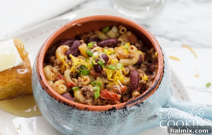 Chili Mac is a delicious combination of ground beef, beans, tomatoes, chili seasoning, and macaroni. This chili mac recipe makes a hearty dinner or a delicious cup of warmth for friends watching football! #chilimac #chili #groundbeef #beans #macaroni #footballrecipes #comfortfood #winterfood #chili A hearty serving of chili mac, accompanied by a slice of warm cornbread drizzled with honey, highlighting the perfect comfort food combination.