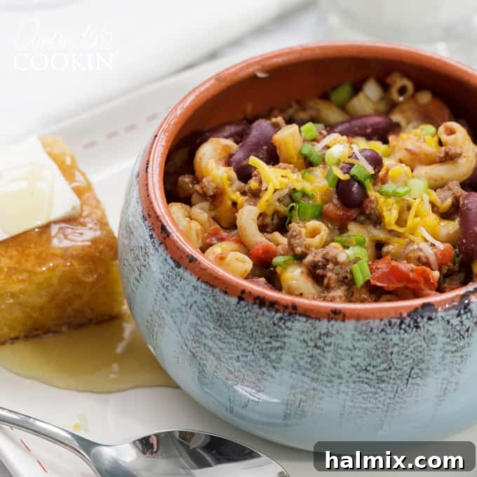Chili Mac is a delicious combination of ground beef, beans, tomatoes, chili seasoning, and macaroni. This chili mac recipe makes a hearty dinner or a delicious cup of warmth for friends watching football! #chilimac #chili #groundbeef #beans #macaroni #footballrecipes #comfortfood #winterfood #chili A close-up shot of a bowl of chili mac, with a side of golden cornbread, emphasizing its inviting texture and deliciousness.