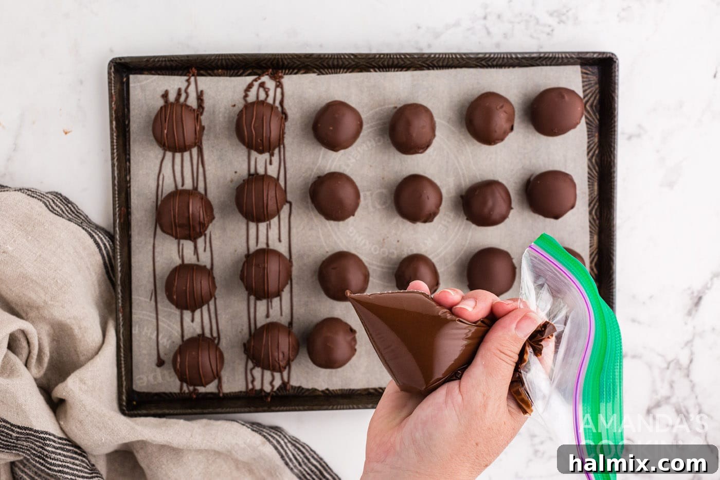 Melted chocolate being drizzled from a piping bag over finished chocolate peanut butter balls on a sheet pan.