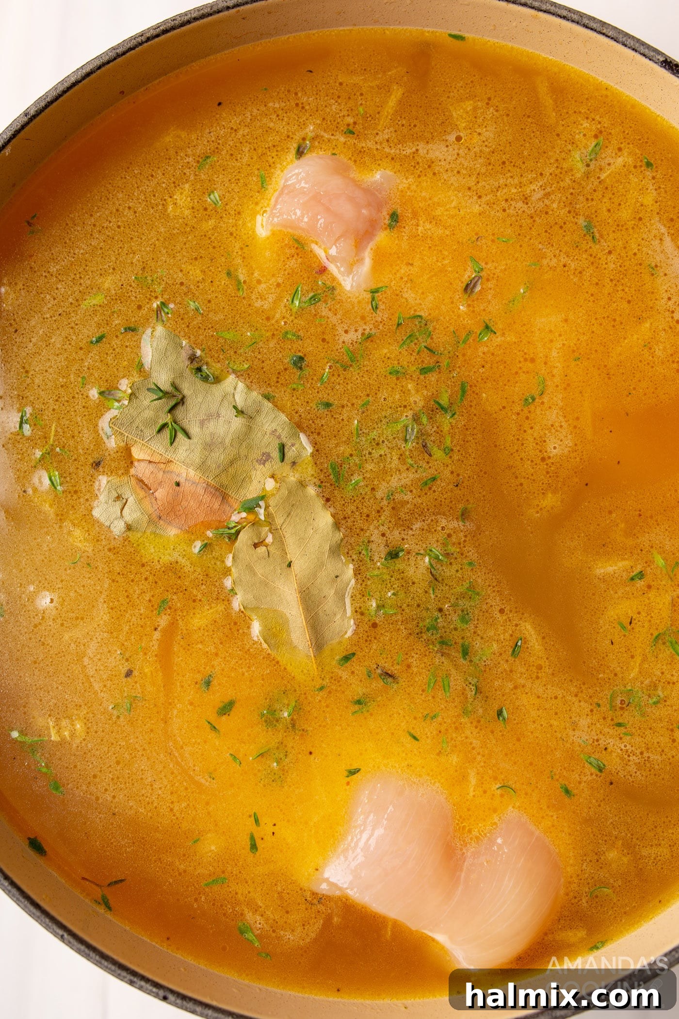 Golden Noodle Chicken Comfort 6 Raw chicken breasts submerged in chicken noodle soup broth with bay leaves and thyme, simmering in a pot.