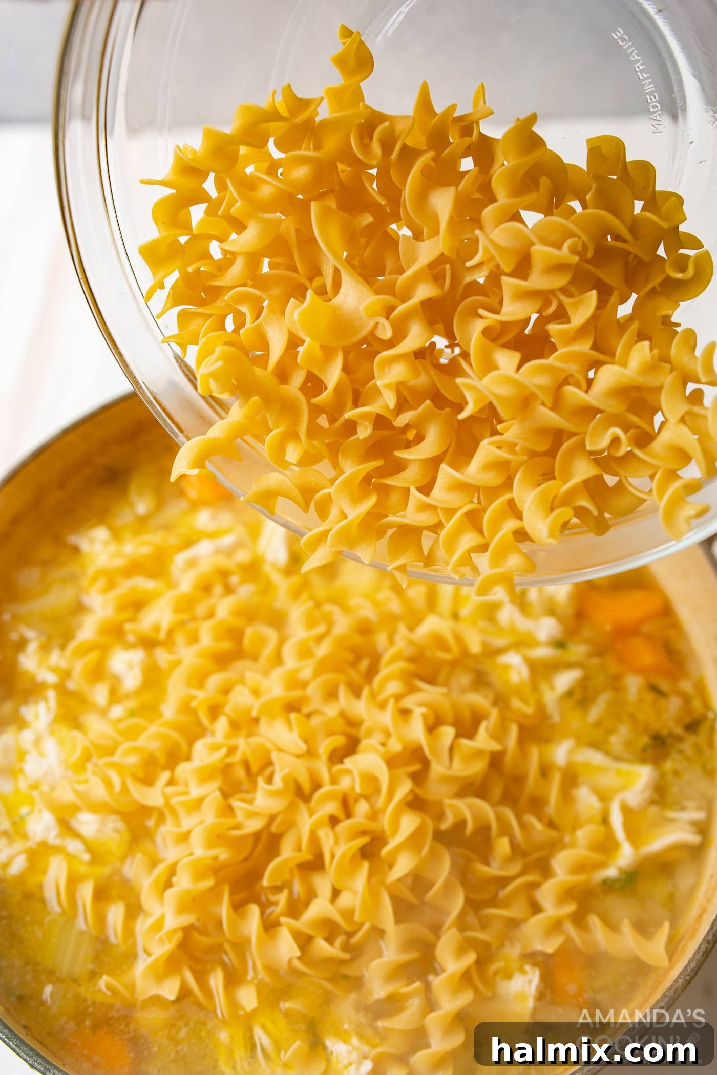 Golden Noodle Chicken Comfort 7 Uncooked egg noodles being poured into a pot of simmering chicken noodle soup broth with shredded chicken.