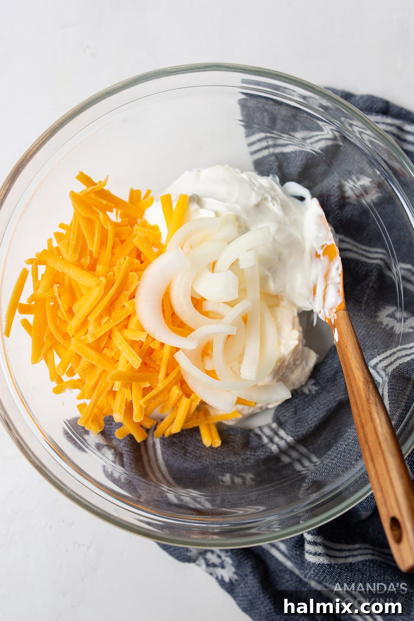 A bowl containing a creamy mixture of sour cream, mayonnaise, half of the cheddar cheese, and half of the onions, stirred and ready for the casserole.