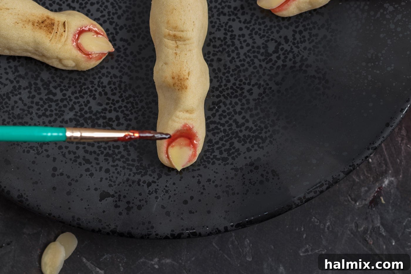 Hexed Hand Delights 15 A hand carefully painting strawberry jam around the almond nail of a baked witch finger cookie, creating a 'bloody' effect.