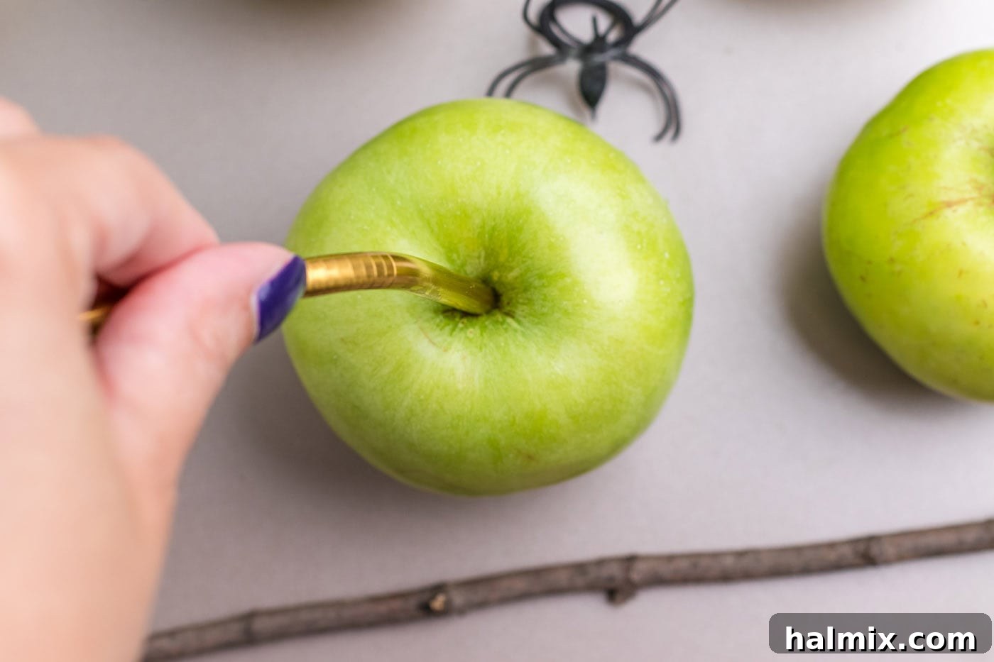 A hand using a metal straw to create a hole in the top of a green apple.