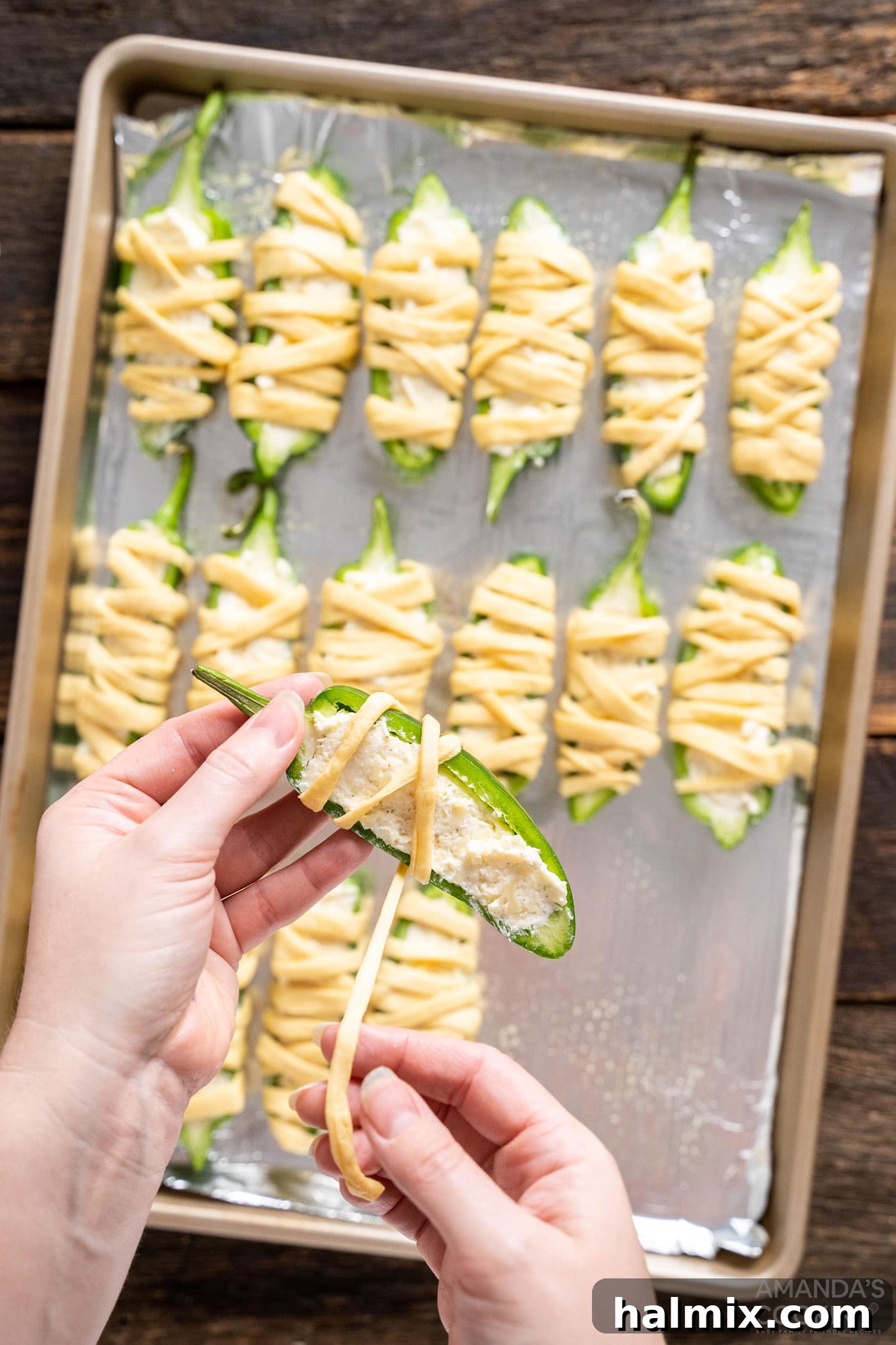 Wrapping stuffed jalapenos with crescent dough strips, leaving a gap for the mummy's eyes
