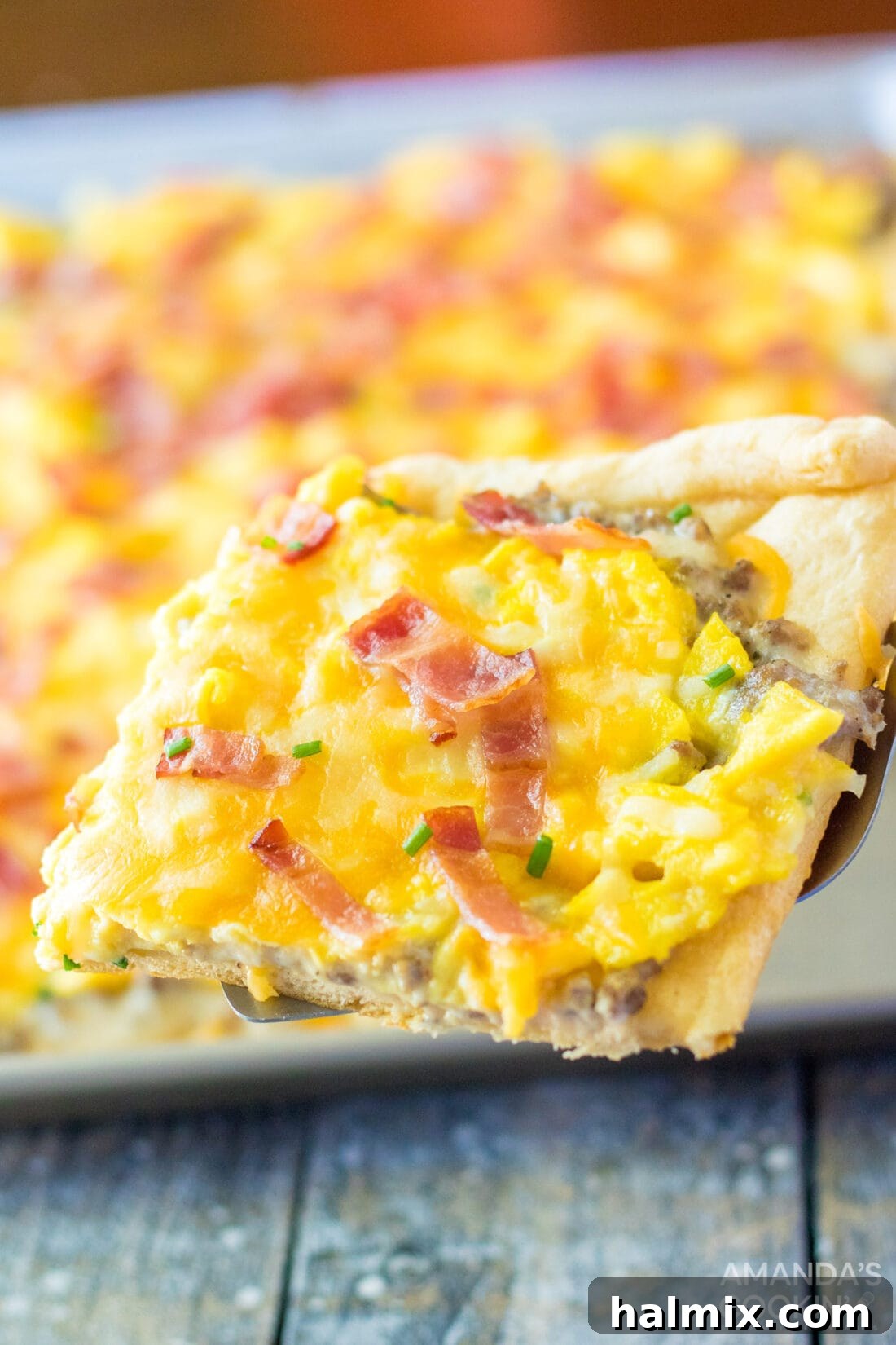A spatula lifting a perfectly golden piece of Sheet Pan Breakfast Pizza, revealing its fluffy crust and layers of eggs, cheese, and sausage gravy.