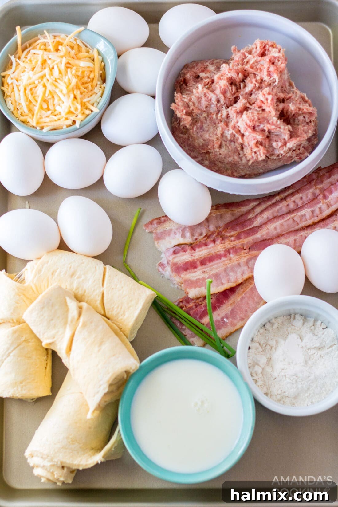 A flat lay photograph showcasing all the fresh ingredients needed for Sheet Pan Breakfast Pizza, including crescent rolls, raw breakfast sausage, eggs, bacon, cheese, milk, and flour.