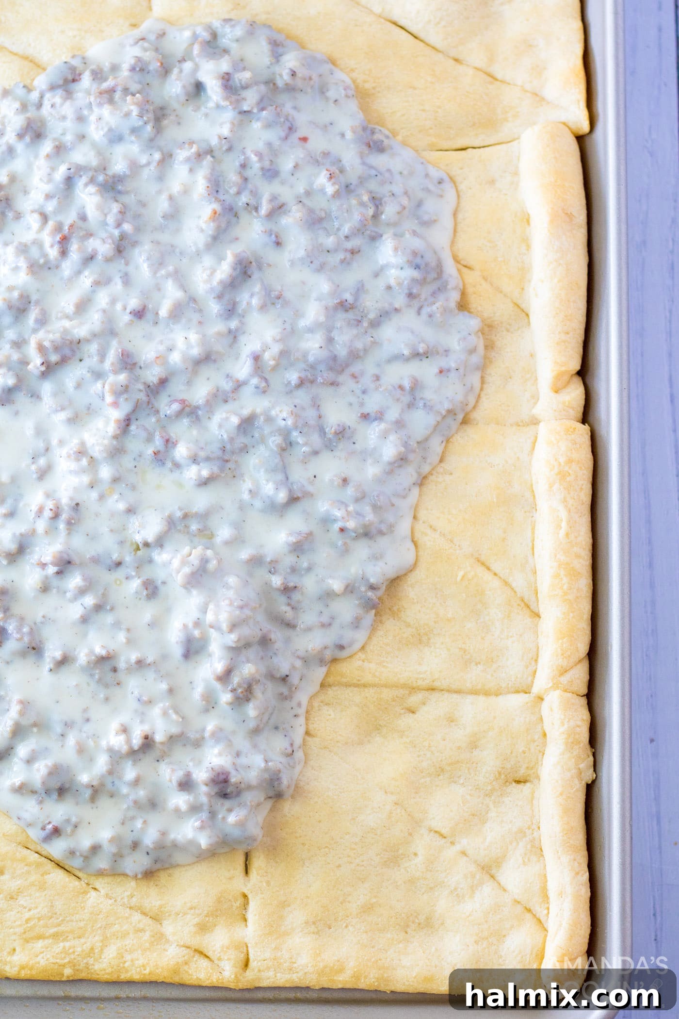 Sausage gravy spread evenly over the golden-brown crescent roll crust on a sheet pan, ready for the next layer of toppings.
