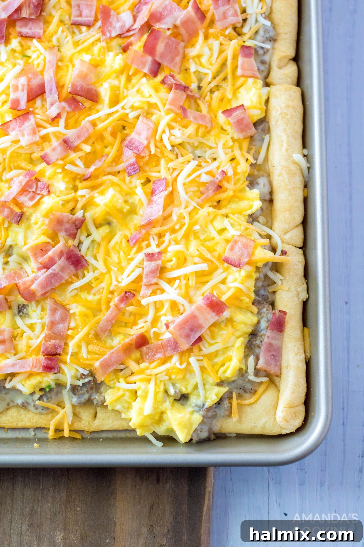 A close-up view of the assembled Sheet Pan Breakfast Pizza, showing layers of shredded cheese, fluffy egg, and crumbled sausage on the crescent dough crust, before the final bake.