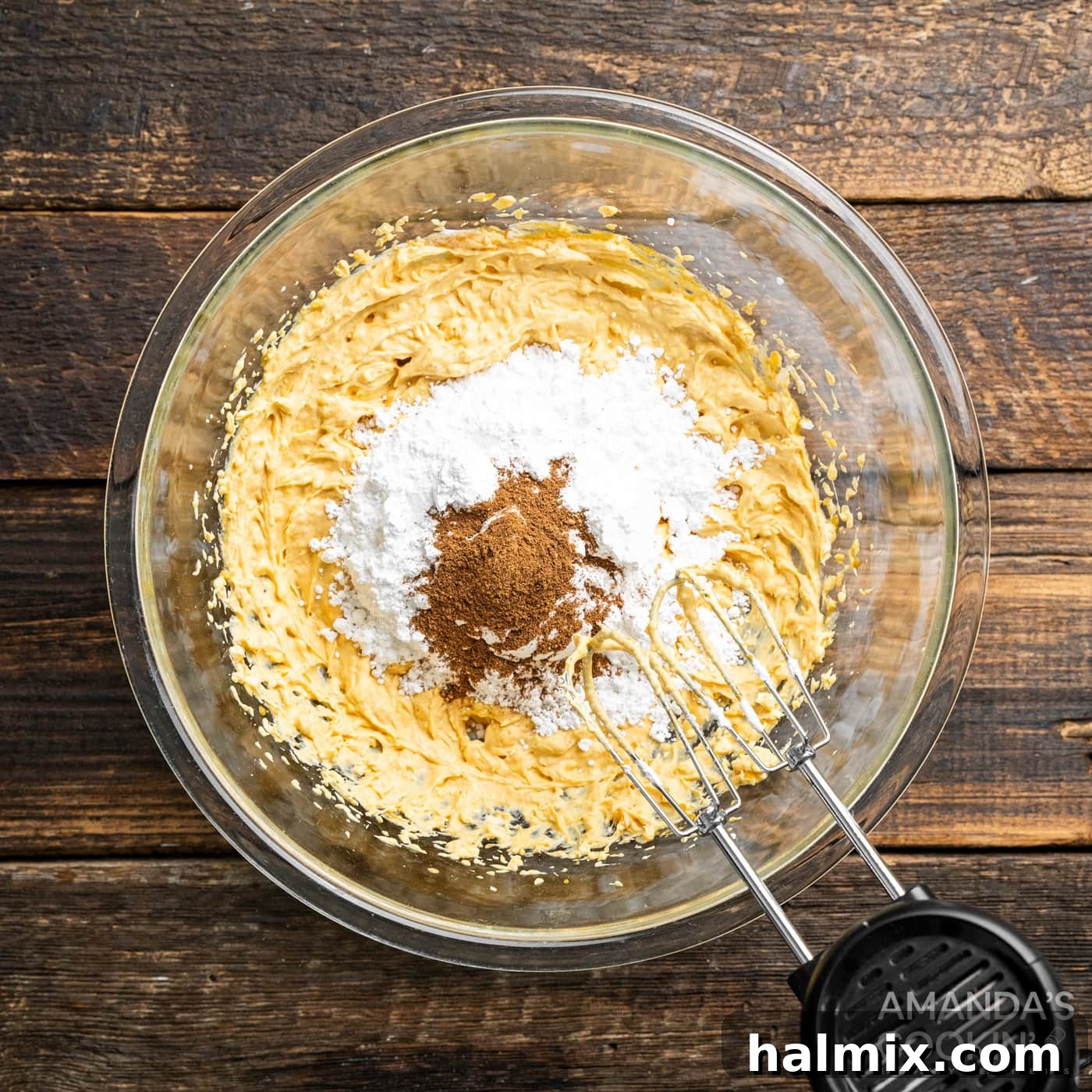 The handheld mixer continues to work, blending powdered sugar, pumpkin pie spice, and vanilla extract into the pumpkin and cream cheese mixture, further developing its rich flavor and smooth texture.