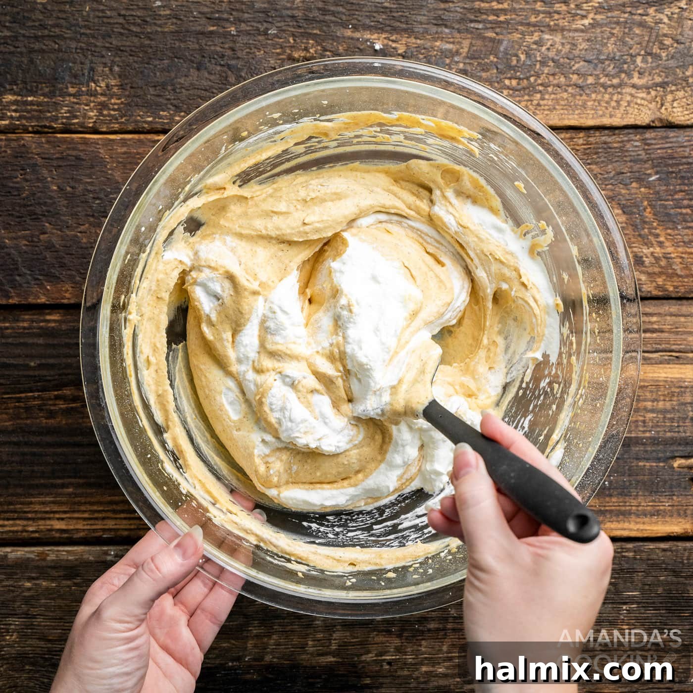 A spatula gently folds the remaining fluffy whipped cream into the light pumpkin and cream cheese mixture, ensuring a smooth and airy consistency without deflating the cream.