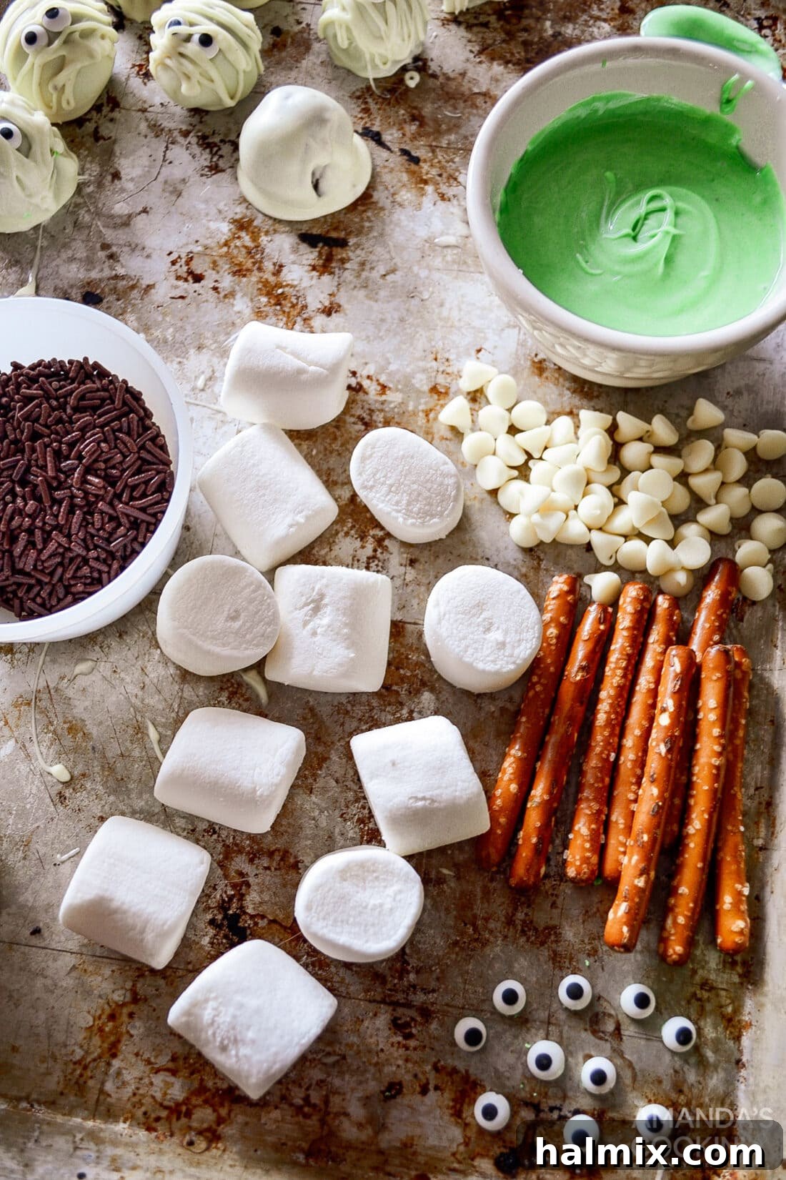 Various ingredients for Marshmallow Frankensteins laid out, including marshmallows, chocolate, sprinkles, candy eyes, and pretzel sticks.