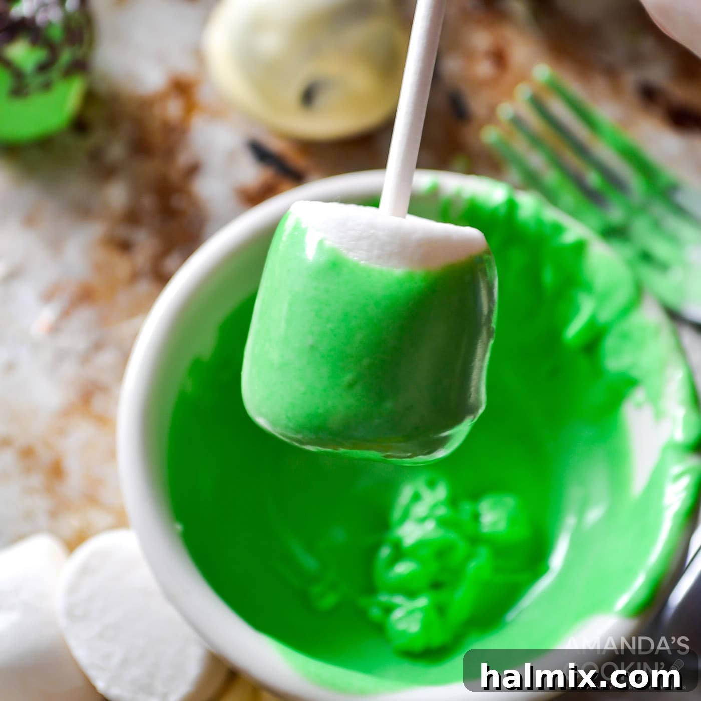 Hand dipping a marshmallow into green chocolate coating, illustrating the first step of decoration.