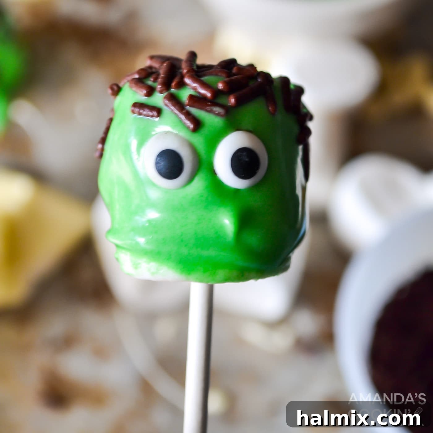 Partially decorated Frankenstein marshmallow pops with green chocolate, sprinkles, and candy eyes, awaiting pretzel bolts.