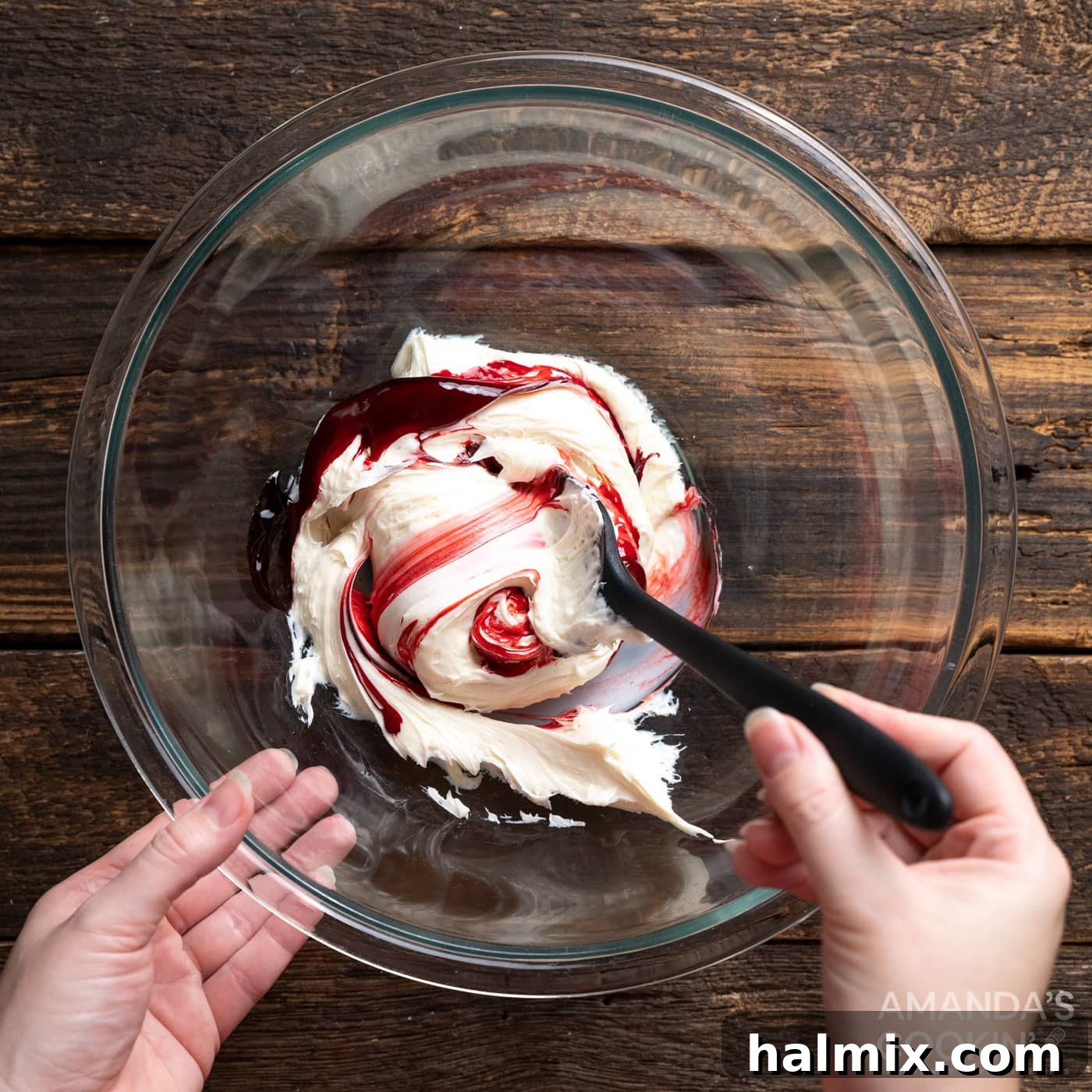 A bowl of white vanilla frosting being mixed with red gel food coloring to create a vibrant red hue.