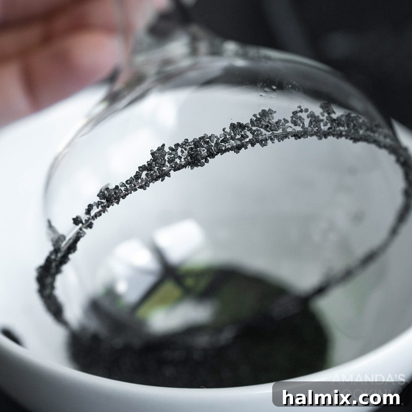A margarita glass being dipped into a shallow plate filled with black lava salt, creating a dramatic rim.