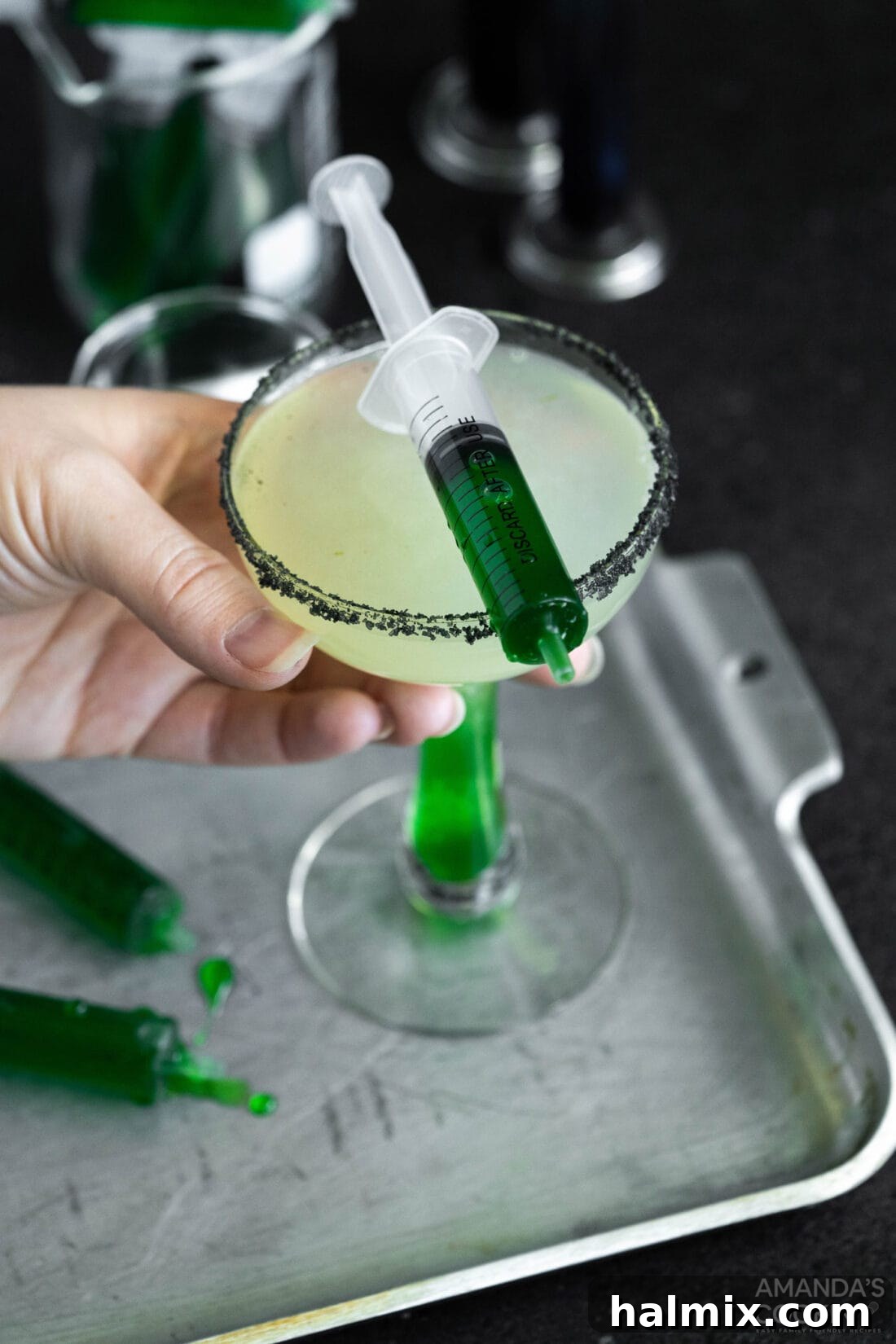 A hand lifting a Frankenstein Margarita, showing off its striking green color, black salt rim, and a lime wedge garnish.