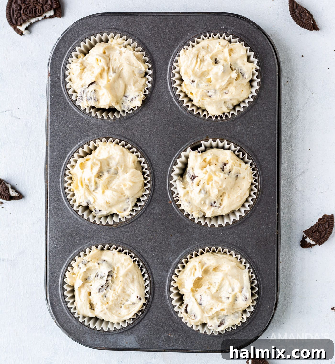 Cupcake batter, mixed with crushed Oreos, has been spooned into paper-lined cupcake cavities, each containing a whole Oreo at the bottom, filling them about three-quarters full.