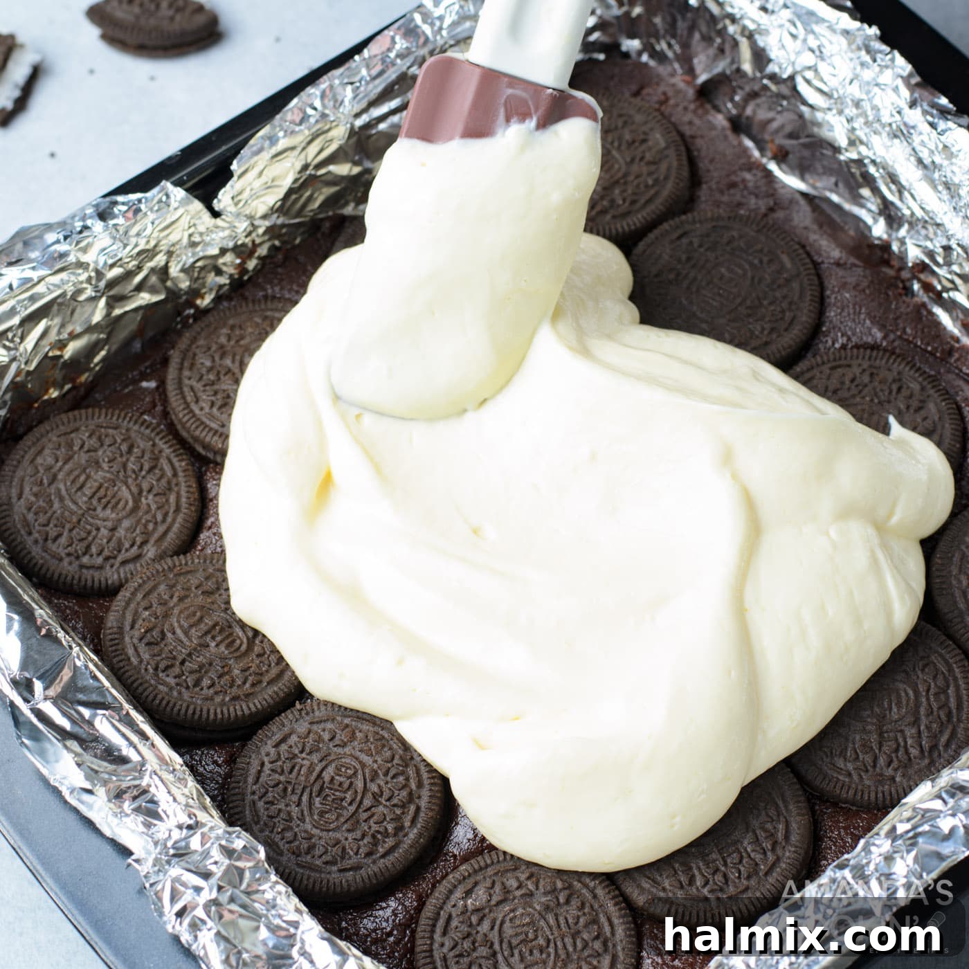 Spatula smoothing out the cheesecake layer on top of the Oreos and brownies